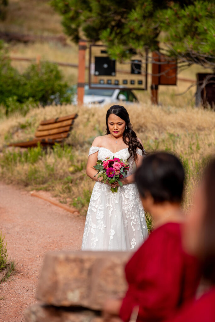 Bride holding a vibrant bouquet walks a gravel path in her lace wedding dress during an outdoor Boulder Halfway House wedding.