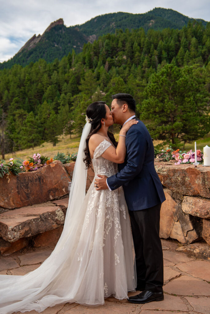 Bride and groom share their first kiss during a Boulder Halfway House wedding ceremony with stunning Flatirons in the background.