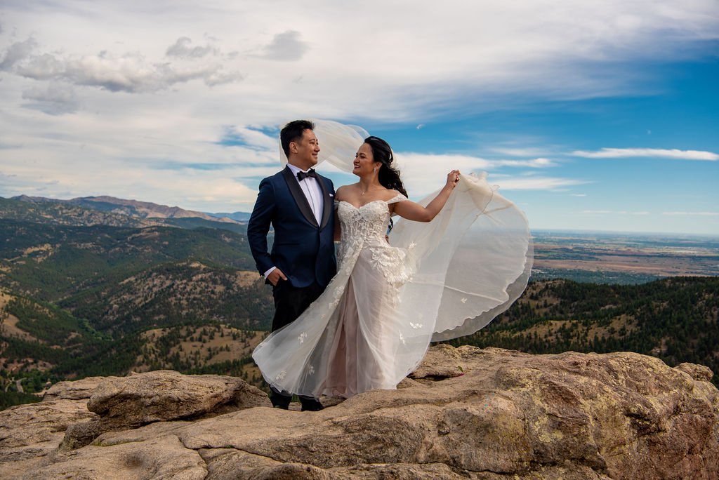 Bride twirls her veil in the breeze while standing with her groom on a mountaintop at their scenic Boulder Halfway House wedding.