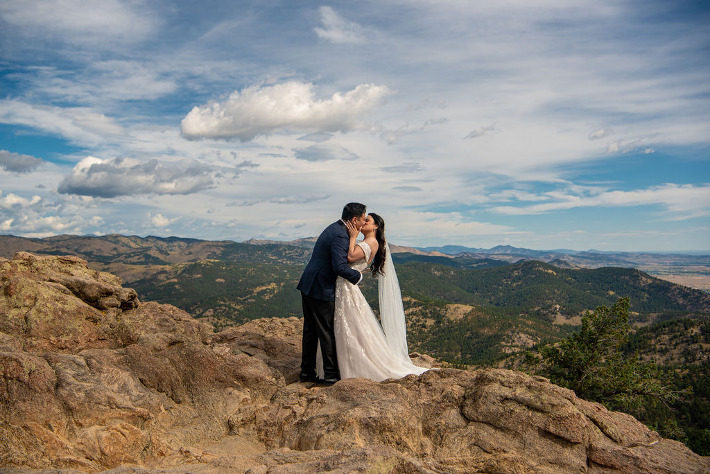 Newlyweds share a romantic kiss atop a rocky overlook with the Colorado mountains as a backdrop at their Boulder Halfway House wedding.