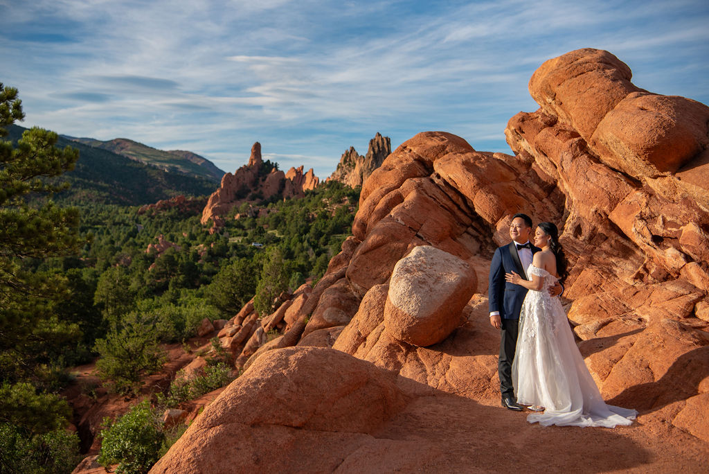 A newlywed couple poses on vibrant red rock formations during golden hour, surrounded by the striking landscape of Garden of the Gods. The bride wears an off-the-shoulder lace wedding dress with a flowing train, and the groom is in a navy blue tuxedo. Towering sandstone spires and forested hills frame the scene under a blue sky with wispy clouds.