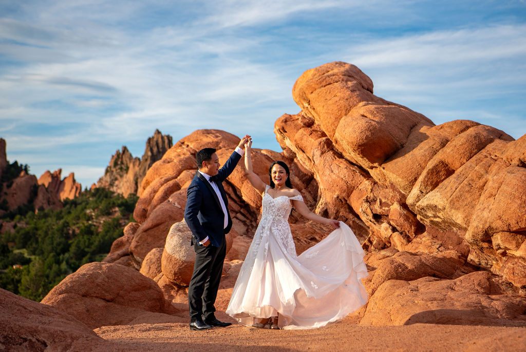The groom twirls his bride in front of towering red rock formations at Garden of the Gods after their wedding. The bride beams, holding her gown as it flows gracefully with her movement. The golden-hour sunlight highlights the rocky landscape, adding warmth and romance to the joyful scene.