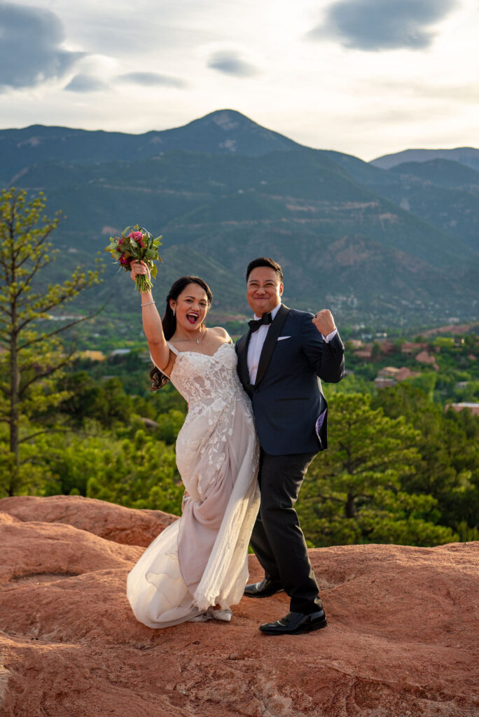 A joyful bride and groom celebrate their wedding, standing on a scenic red rock ledge with lush green trees below and towering mountains in the distance. The bride raises her bouquet triumphantly, wearing a flowing white lace gown, while the groom in a navy tuxedo fist-pumps beside her under a golden sunset sky.
