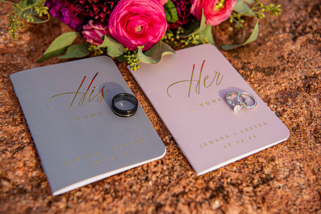 Close-up of two vow booklets labeled “His Vows” and “Her Vows” placed on a rocky surface. The “His” booklet is slate gray with a black wedding band on top, while the “Her” booklet is light blush with two silver rings, including a diamond engagement ring. Behind them, a bouquet of vibrant flowers, including pink ranunculus and eucalyptus, adds a pop of color.