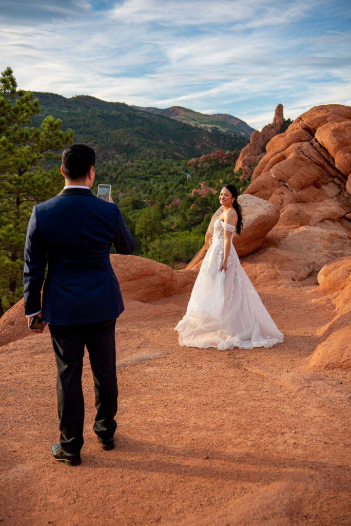 The groom takes a smartphone photo of his bride, who smiles confidently while posing on a rocky trail. Her elegant gown stands out against the red terrain and green trees below the mountain ridges. The light of sunset adds a romantic glow to the scene. Boulder Halfway House wedding portrait session overlooking Colorado’s red rock formations.