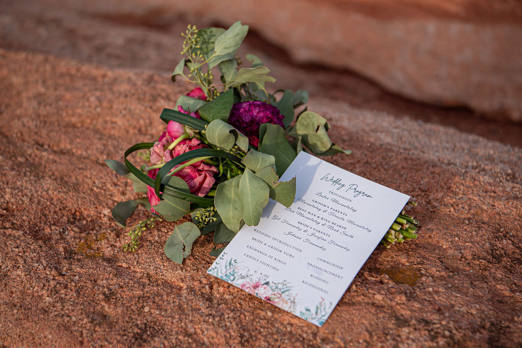 A floral bouquet featuring pink and purple blooms with eucalyptus greenery rests on red rock beside a printed wedding program. The program lists traditional ceremony elements such as processional, exchange of vows, and ring exchange, all framed with soft floral illustrations.