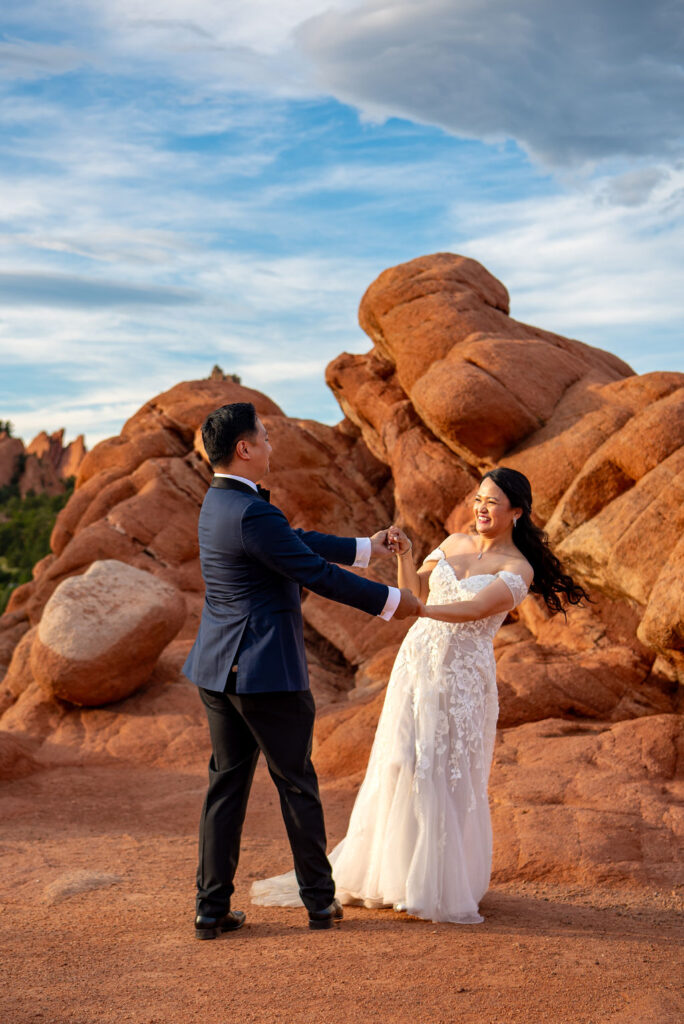 The couple shares a joyful moment dancing on a red dirt path at the base of large rock formations. The bride’s white lace dress sways as she laughs, holding hands with her groom in a navy suit. Behind them, the sun casts warm light on the textured rocks and distant greenery. Playful moment captured in the Colorado foothills.