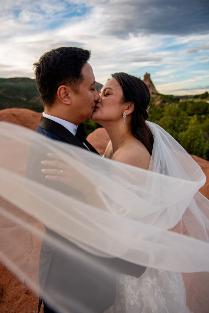 Close-up of the bride and groom kissing tenderly, their faces framed by the bride’s sheer white veil flowing in the wind. The bride’s delicate earrings and soft makeup shine in the natural light, while the mountains and rocks of the Colorado landscape blur in the background. Intimate kiss at a Boulder Halfway House wedding with scenic mountain backdrop.