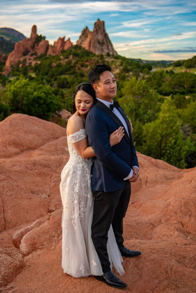 A romantic moment during
this couple's wedding shows the bride gently embracing the groom from behind as they stand on red rocks at Garden of the Gods. The bride closes her eyes, resting her head on the groom’s back, while he gazes ahead in a navy suit. Iconic rock spires and greenery fill the dramatic background.