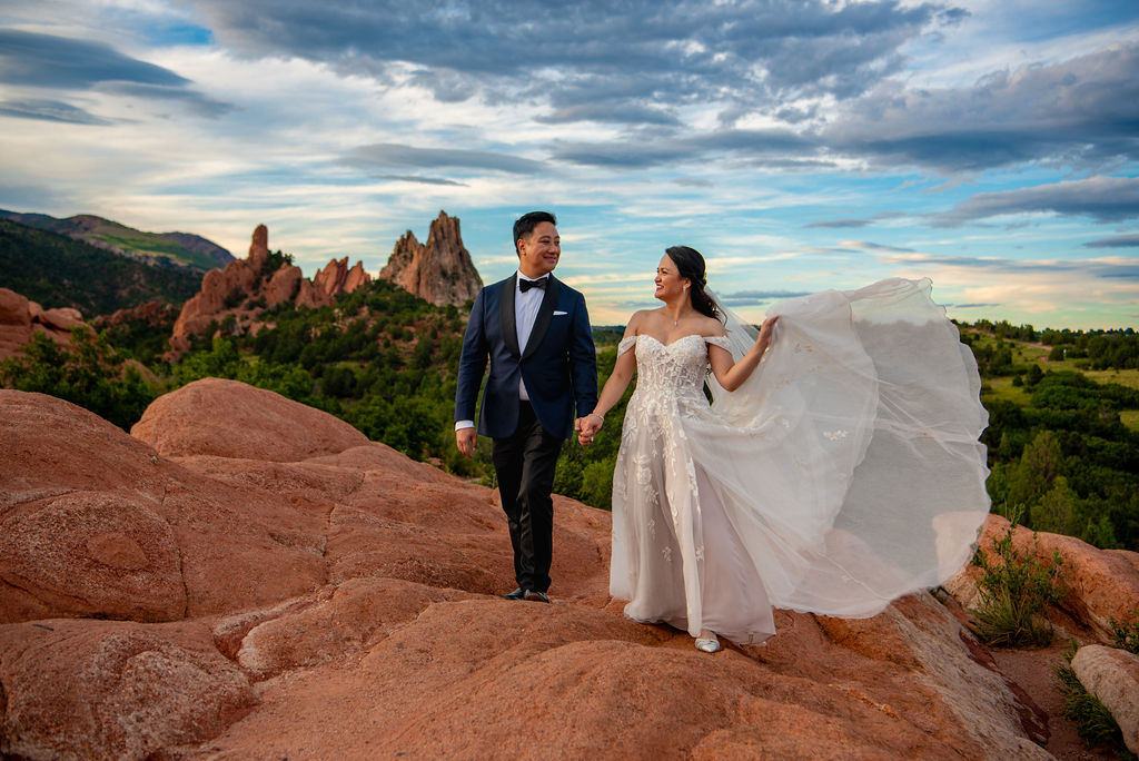 A bride and groom walk hand in hand across striking red rock formations with dramatic spires rising behind them at sunset. The bride’s white gown billows gracefully in the breeze, creating a sense of movement and freedom. She holds her skirt with one hand and gazes at the groom, who is smiling back at her while wearing a formal navy tuxedo. The rich orange and red tones of the rocky terrain contrast beautifully with the deep greens of the surrounding vegetation and the vivid blues and purples in the dramatic, cloud-streaked sky.