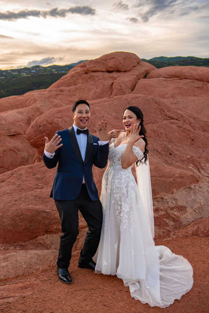 The newlyweds share a playful moment during their wedding, excitedly showing off their wedding rings. They stand against striking red rock formations at Garden of the Gods, the bride in a lace strapless gown and the groom in a navy tuxedo. Both wear huge smiles, framed by a dramatic, colorful sky.