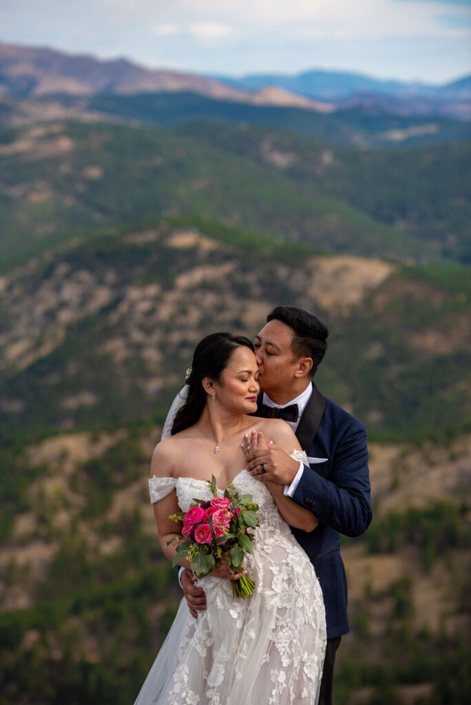 The groom tenderly kisses the bride on the temple as they embrace in front of a vast mountain landscape. The bride closes her eyes and smiles softly, holding a bouquet of deep pink roses and green foliage. She wears an elegant, lace off-the-shoulder wedding dress and a delicate necklace, while the groom, in a navy suit and black bow tie, lovingly wraps his arms around her from behind. The scene is serene and romantic, with rolling forested mountains fading into the distance under a softly clouded sky.