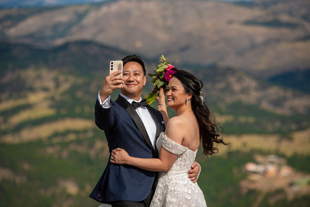 A joyful bride and groom take a selfie on their wedding day against a breathtaking mountain backdrop. The groom, wearing a dark navy tuxedo with a black lapel and bow tie, holds a smartphone in his outstretched hand while smiling at the camera. The bride, dressed in a white off-the-shoulder lace wedding gown, stands beside him with her arm around his waist, smiling brightly. She holds a bouquet of vibrant pink and red flowers with greenery. The background reveals sunlit green hills and rugged mountain terrain, softened by the warm, golden light.
