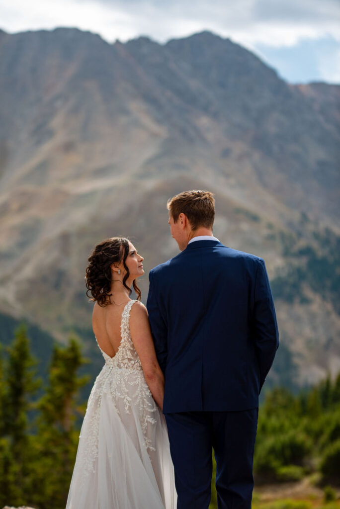 Bride and groom stand hand in hand, facing a majestic mountain backdrop. The bride wears a white lace dress, and the groom is in a navy suit.