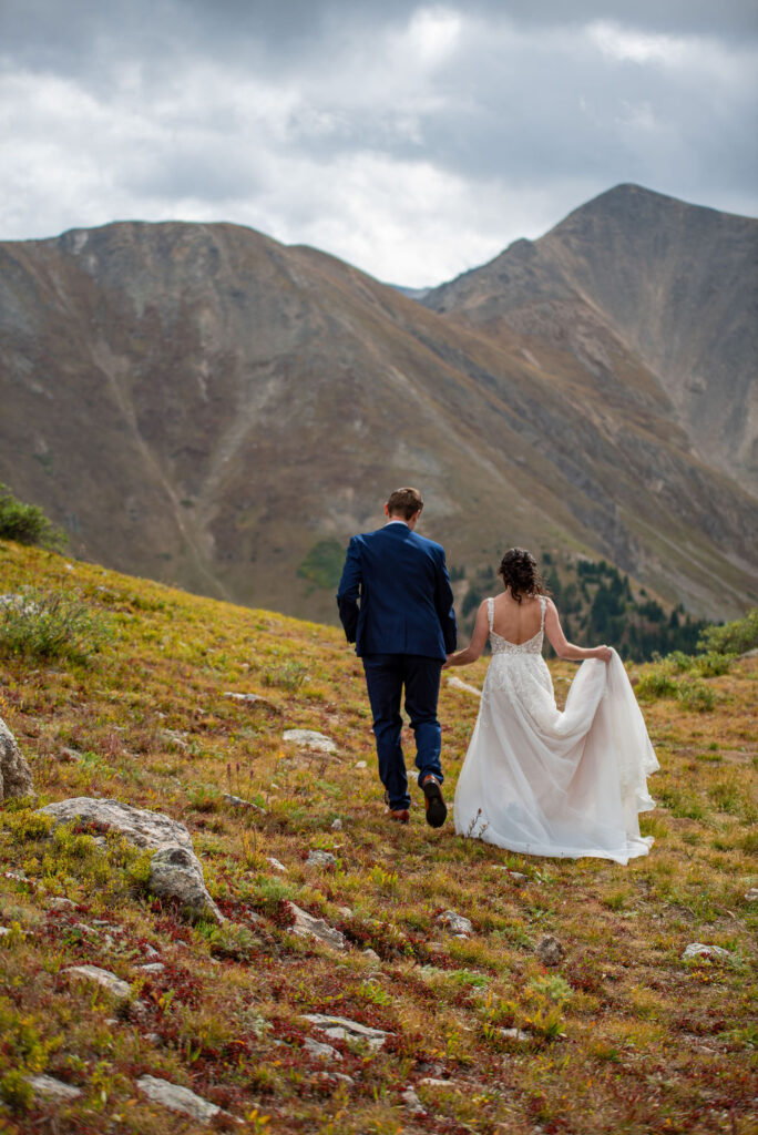 A couple in wedding attire walks hand-in-hand up a grassy hillside, framed by towering mountains under a cloudy sky, evoking romance and adventure. All because their Colorado elopement planning was done carefully.