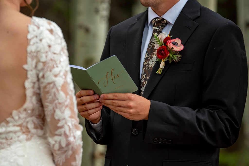 A groom in a dark blue suit holds a "his vows" booklet outdoors. Nearby, pale orange and white flowers add a touch of elegance and romance.
