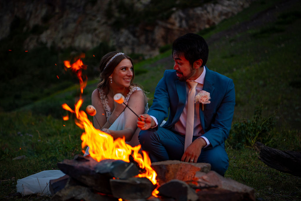 A couple in wedding attire toasts marshmallows over a campfire, set in a grassy mountain landscape. They gaze at each other, smiling warmly. Their Colorado elopement planning ensured they had s'mores by a campfire at the end of their wedding dya.