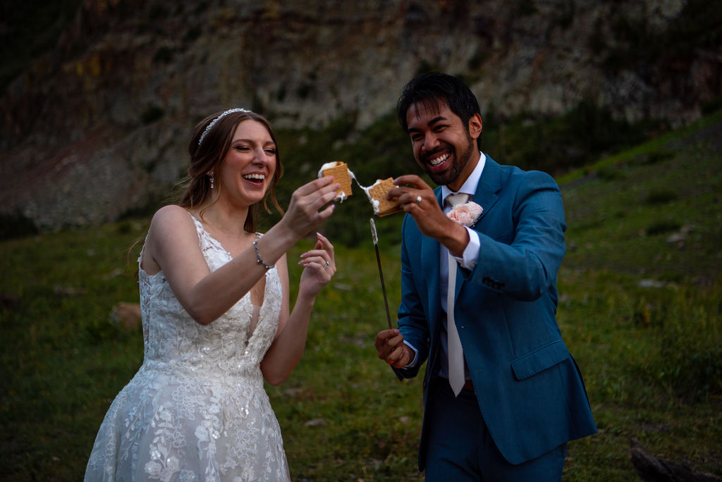 A bride in a lace dress and a groom in a blue suit joyfully toast marshmallows together outdoors, with a rocky hillside in the background. The perfect Colorado elopement planning