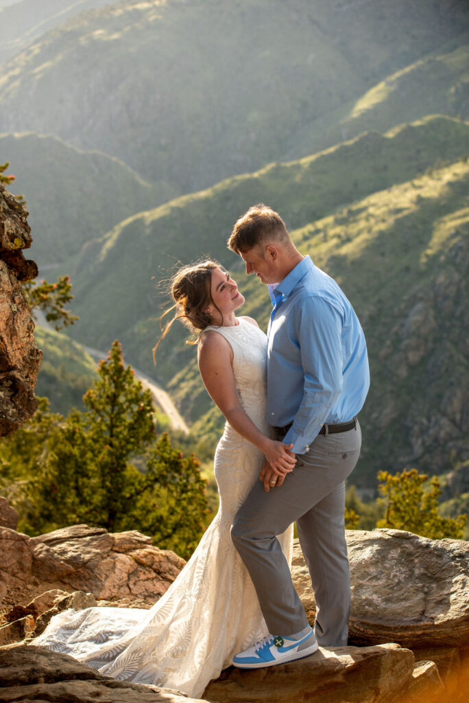 Bride and groom stand on rocky cliff, smiling at each other. Bride in lace dress, groom in blue shirt and sneakers. Lush green mountains behind.