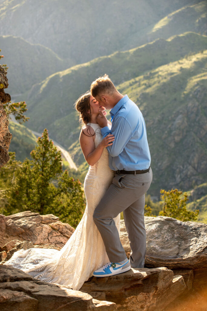 A couple kisses tenderly atop rocky cliffs, with mountains and trees in the background. The woman wears a white dress; the man, a light blue shirt and sneakers. Everything came together for their Colorado elopement planning.