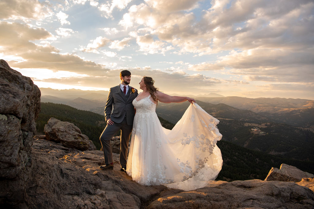A couple poses on a rocky mountain top at sunset; the bride holds her flowing gown. The dramatic sky and distant hills create a romantic, serene atmosphere.
