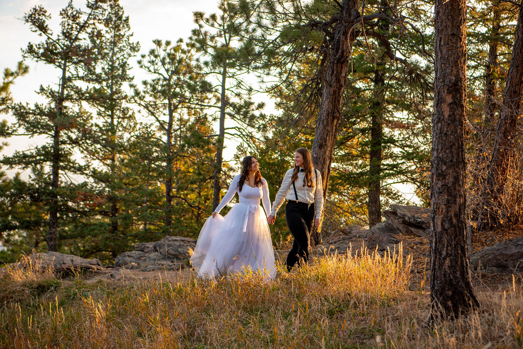 A couple walks hand-in-hand in a forest at sunset. One wears a white wedding dress, the other a shirt and suspenders. Warm light filters through the pine trees.