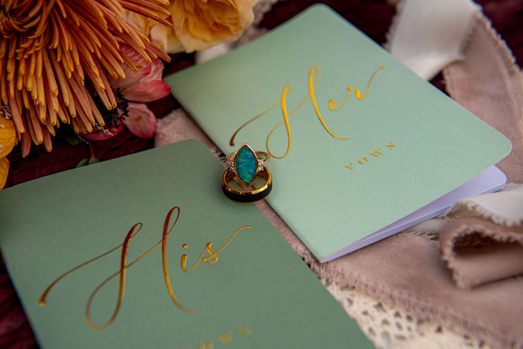 Two vow booklets labeled "His" and "Her" in elegant gold lettering lie beside vibrant flowers and lace. A turquoise ring rests on the "Her" booklet, creating a romantic and intimate atmosphere.