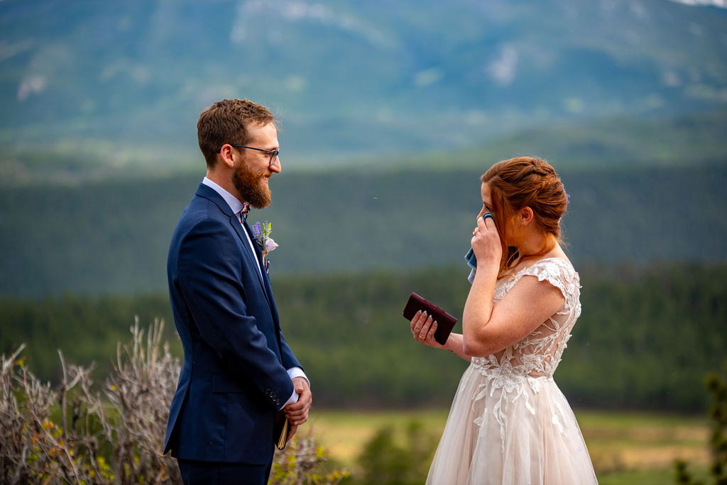 A bride and groom share an emotional moment outdoors. The bride wipes her tears while holding a notebook, as the groom smiles tenderly. Mountain scenery in the background. Their Colorado elopement planning ensured they shared a private ceremony filled with emotions.