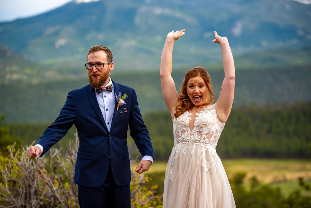 A joyful bride in a lace gown raises her arms in excitement beside the groom, who wears a blue suit. They're outdoors with mountains in the background.