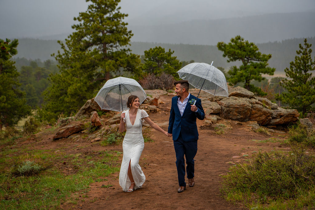 A bride in a white dress and a groom in a blue suit walk hand in hand on a rocky path under clear umbrellas, surrounded by lush, misty forests. All their Colorado elopement planning prepared them for the day filled with rain and it was still perfect.