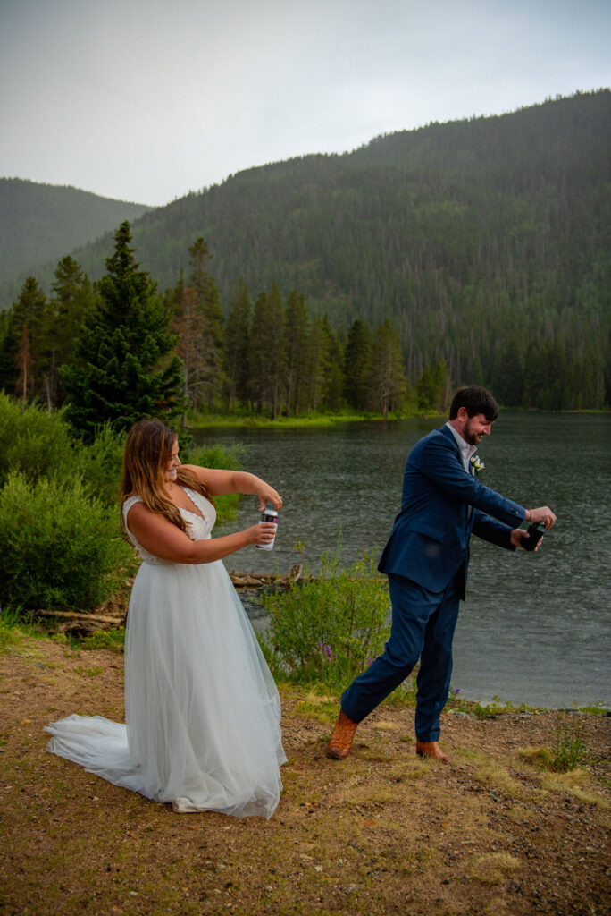 Bride and groom in formal attire joyfully shake drinks and get ready to shot gun a few beers near a serene mountain lake. Lush greenery and forested hills create a tranquil backdrop. They are celebrating their successful Colorado elopement planning.