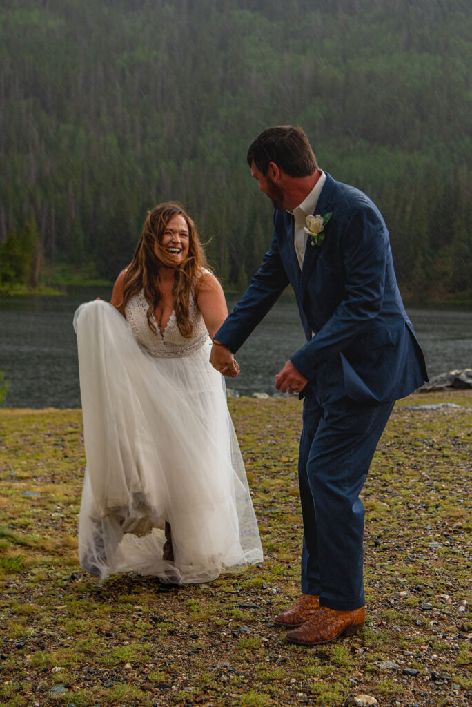A joyful bride and groom laugh under light rain by a forested lake. She holds her white dress, and he wears a blue suit with a boutonniere. They are smiling at each other. Their Colorado elopement planning had paid off.