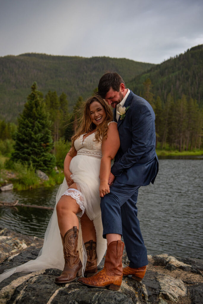 A joyful bride and groom stand on rocks by a lake, wearing cowboy boots. The bride lifts her dress, revealing lace garters. Mountain landscape in the background.