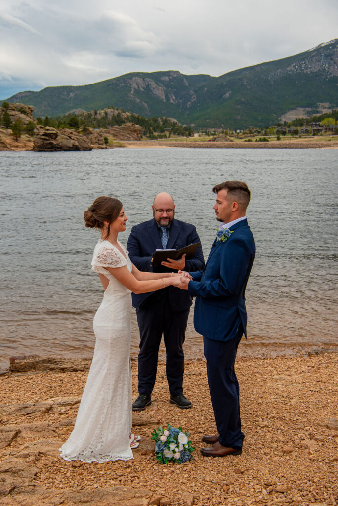 Bride and groom hold hands in front of the officiant as he leads the couple through their wedding ceremony. They are standing in front of a blue lake in Estes Park Colorado. The Estes Park elopement photographer captures the moment before the couple is officially married.