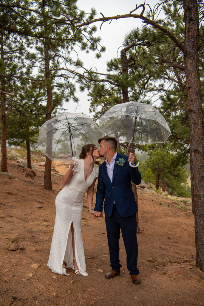 Bride and groom kiss underneath clear umbrellas while standing in a pine forest in a rain storm. The Estes Park elopement photographer captured a quiet moment as the sky is covered in grey clouds and the trail winds behind them.