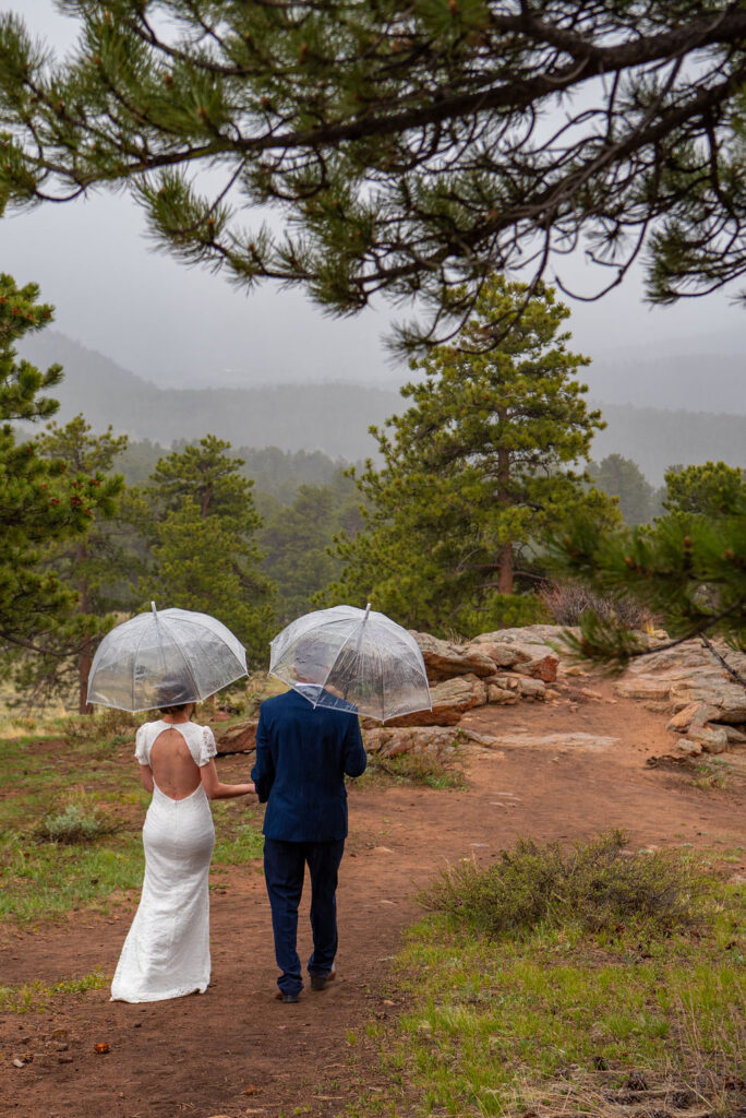 Estes Park elopement photographer captures a moment as the bride and groom hold hands and walk to a clearing in the pine forest. They are holding clear umbrellas to hide from the rain. There is mist and cloud coverage in the background.