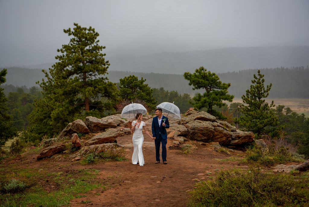 Bride and groom walking hand-in-hand under clear umbrellas along a rocky trail in the mountains during their rainy elopement. Captured by an Estes Park elopement photographer in Rocky Mountain National Park, showcasing romantic moments in unpredictable Colorado weather.
