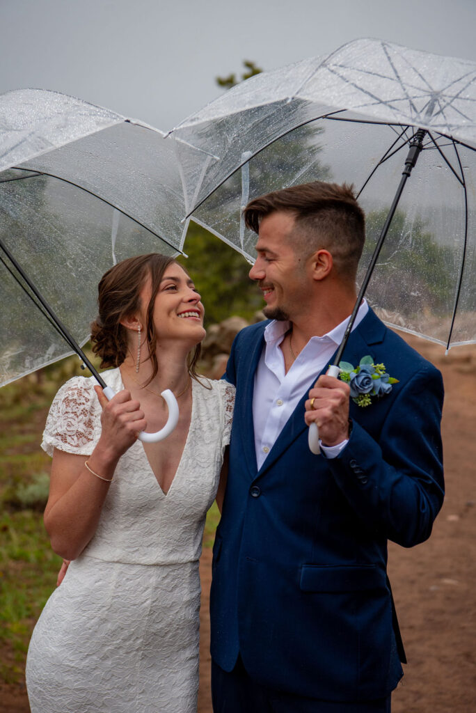 The Estes Park elopement photographer captures a sweet moment a bride and groom as the laugh facing each other while holding clear umbrellas to stay dry from the rain falling from a grey sky.