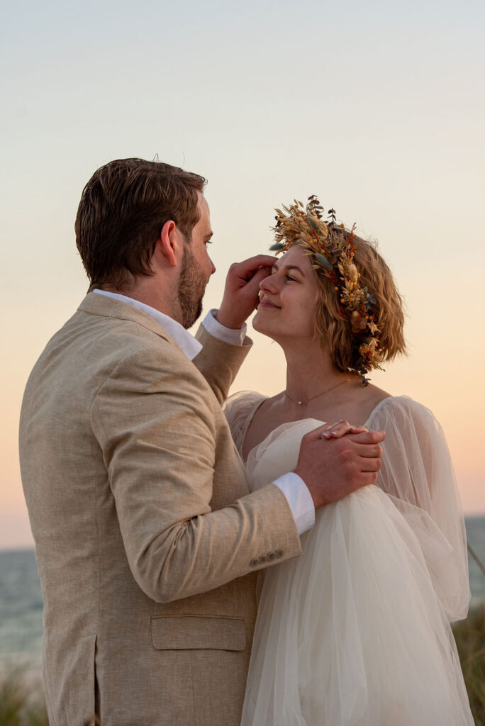 A romantic moment during a National Park elopement on the beach, as a groom gently brushes the bride’s face while they hold each other close at sunset. The bride wears a tulle gown with sheer sleeves and a floral crown, while the groom is dressed in a light tan suit.