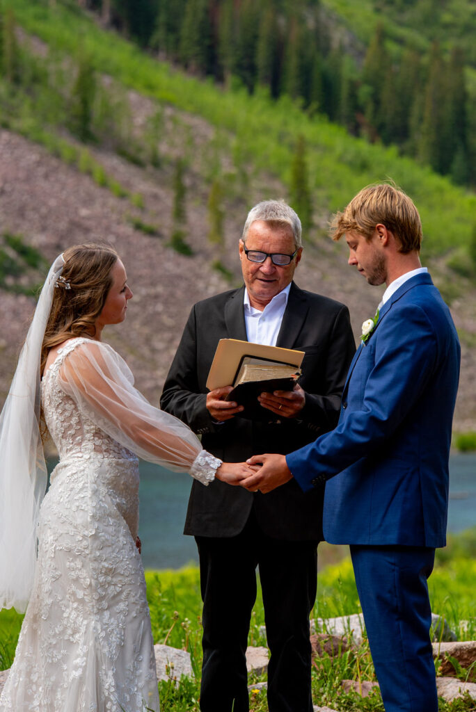 A man and woman holding hands, both are dressed in formal wedding attire. The woman is wearing a wedding dress and veil, while the man is in a suit. They are outdoors on grass with mountains in the background, and they have an officiant standing behind them reading from a book. The perfect example of how to plan an elopement ceremony with an officiant.