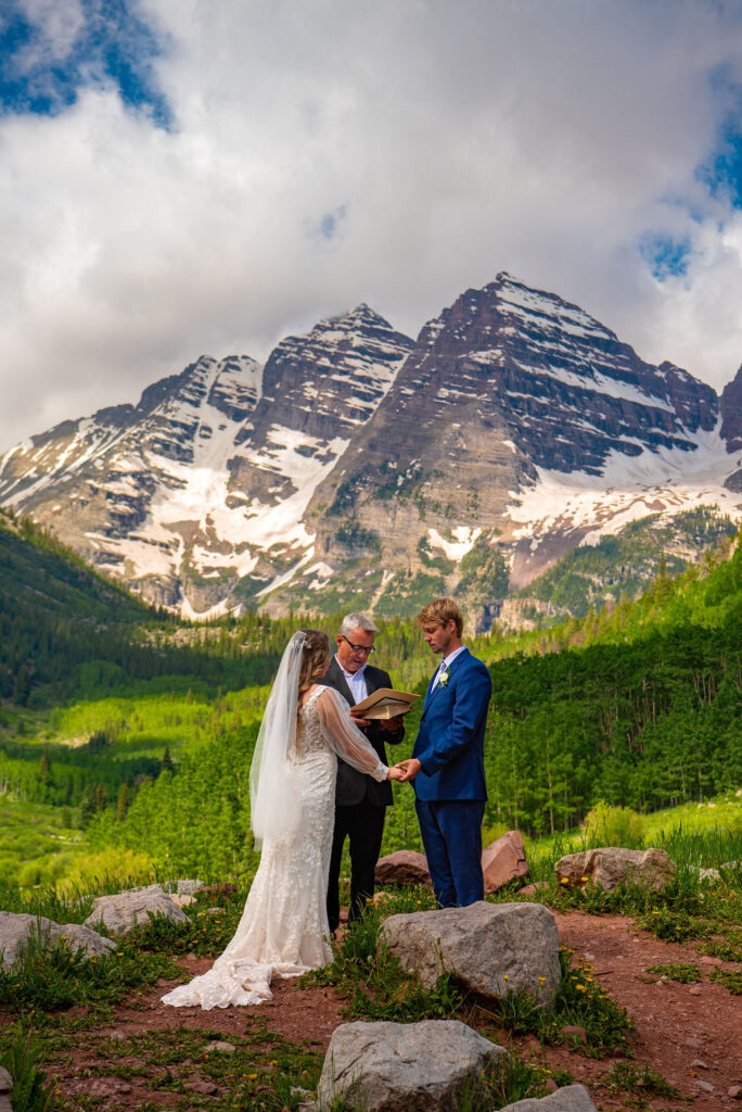 A couple stands together in front of a mountain backdrop, the bride wearing a wedding dress and the groom in formal attire. They are surrounded by grass, with a cloudy sky overhead at the Maroon Bells in Aspen Colorado.