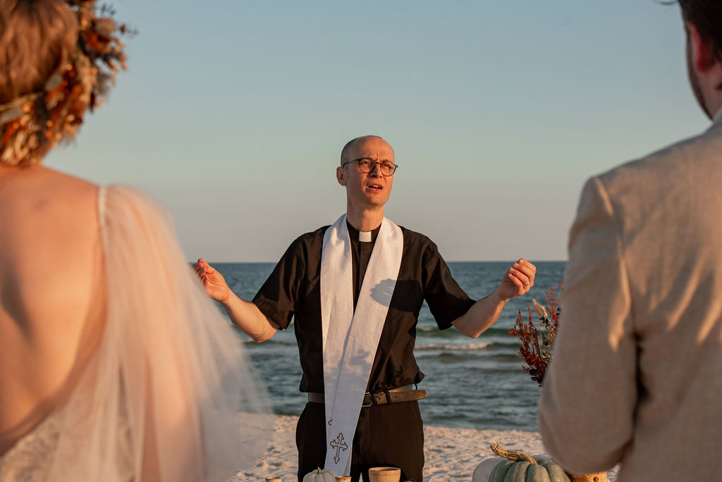 A man wearing a black shirt and a white sash stands on the beach, with a bride and groom in the foreground. The man in black is officiating the wedding ceremony.