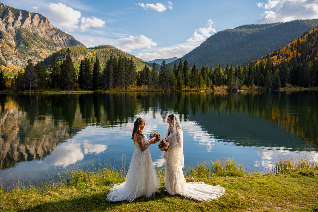 Two women in white dresses stand by a serene lake, surrounded by lush grass and trees. The backdrop features mountains under a partly cloudy sky, adding a picturesque element and all the inspiration on how to plan an elopement ceremony.