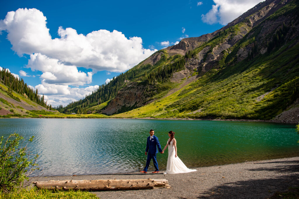 A couple stands hand in hand by a serene, turquoise lake surrounded by lush green mountains under a bright blue sky with fluffy clouds. The couple is wearing wedding attire. The man has a blue suit on and the woman is wearing a lace dress.