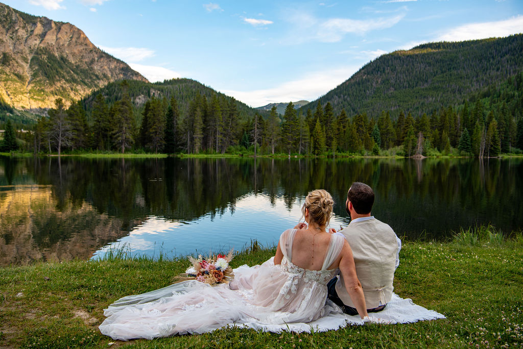 A couple sits on a picnic blanket in the grass by a serene lake, dressed in wedding attire, with mountains and trees reflected in the water. This spring elopement was one of the best times for an elopement in Colorado.