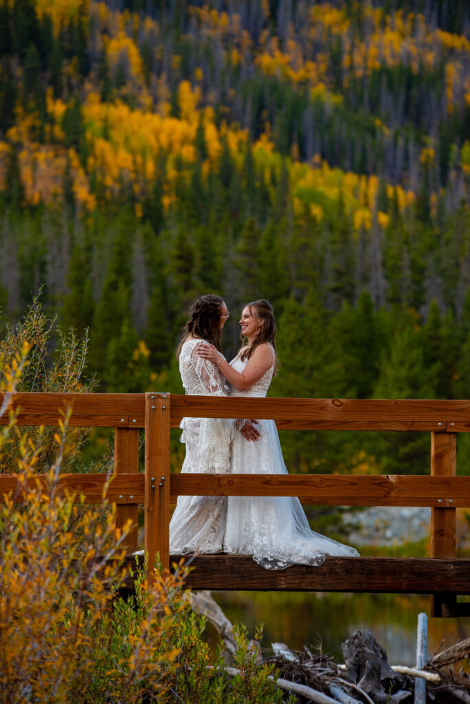 Two brides stand on a wooden bridge, surrounded by vibrant autumn foliage reflecting on a serene lake showing by fall is one of the best times to elope in Colorado.