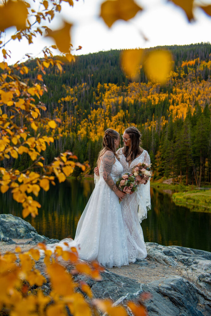 Two brides in elegant white dresses embrace on a rock by a serene lake, surrounded by vibrant autumn foliage which is one of the best times to elope in Colorado.
