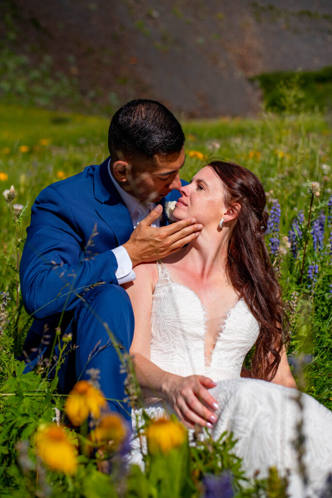 A couple sits in a vibrant flower field, wearing a wedding suit and a delicate lace dress, sharing a tender moment together. Surrounded by different colored wildflowers, this shows why summer is the best time to elope in Colorado.