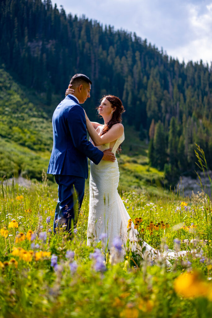 A couple in formal wedding attire embrace amidst a vibrant field of wildflowers, set against a backdrop of lush green mountains in the state of Colorado. Really one of the best times to elope.