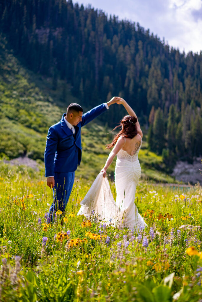 A couple dances in a vibrant flower-filled meadow, surrounded by lush greenery and mountains under a bright sky. This scene shows why summer is the best time to elope in Colorado.